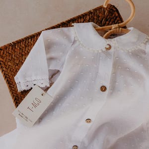 以下が含まれることがあります： ピーターパンカラーと木製ボタンが付いた白いベビー服。長袖シャツは袖口にレーストリムが付いています。この衣装は、木製のハンガーに掛けられ、編まれたバスケットの中にあります。「T-AGO」と書かれたタグが付いています。