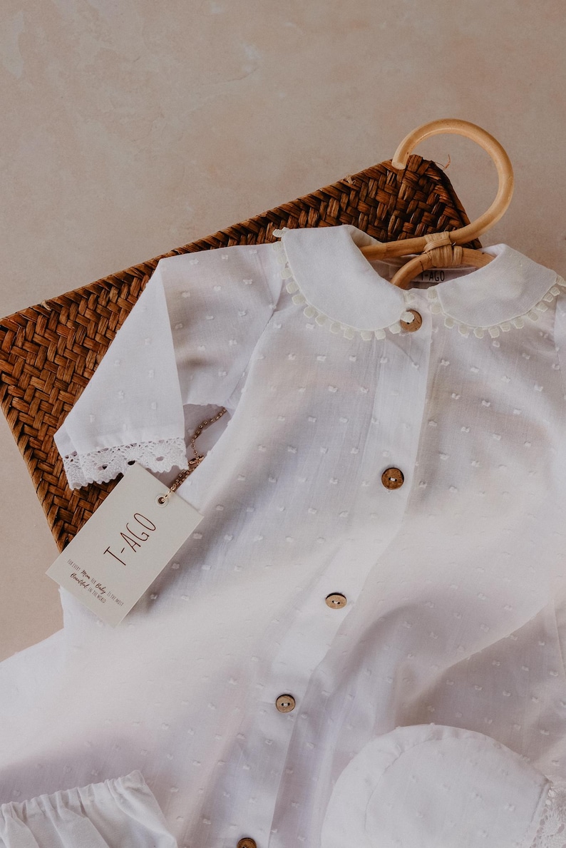 以下が含まれることがあります： ピーターパンカラーと木製ボタンが付いた白いベビー服。長袖シャツは袖口にレーストリムが付いています。この衣装は、木製のハンガーに掛けられ、編まれたバスケットの中にあります。「T-AGO」と書かれたタグが付いています。