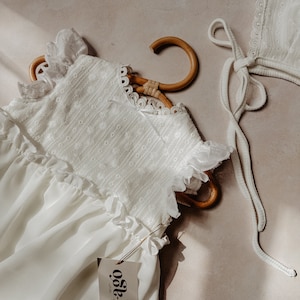 May include: A white baby dress with frilled sleeves and a matching bonnet. The dress is suspended on a wooden hanger. The dress and bonnet are made of a delicate fabric with embroidered details. A tag with the brand name "t-ago" is attached to the dress.