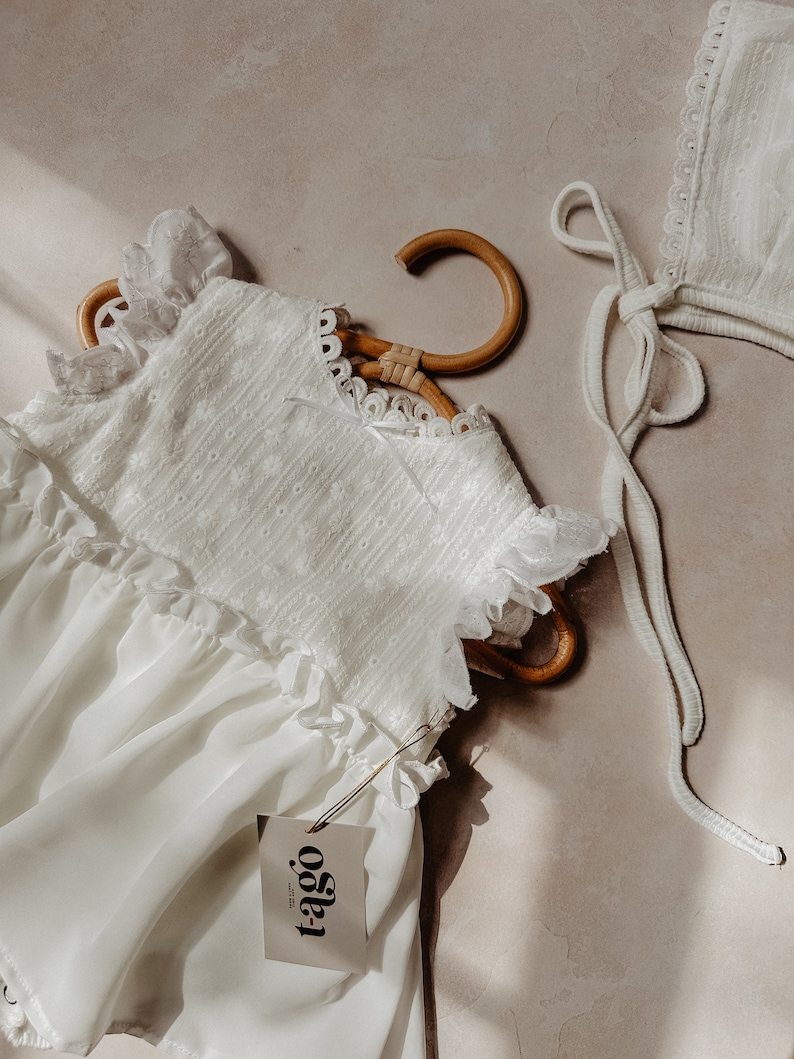 May include: A white baby dress with frilled sleeves and a matching bonnet. The dress is suspended on a wooden hanger. The dress and bonnet are made of a delicate fabric with embroidered details. A tag with the brand name "t-ago" is attached to the dress.