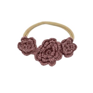 May include: A mauve crochet headband featuring three flower accents. The flowers are made of a textured yarn and are attached to a neutral-colored elastic band. This handmade accessory is suitable for babies and young children.