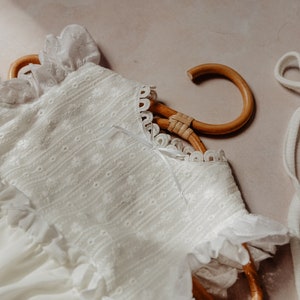 May include: A white baby dress with floral embroidery and ruffled trim hangs on a wooden hanger. The dress features a delicate lace collar and a small bow. The soft, light-coloured fabric suggests a vintage or heirloom style.