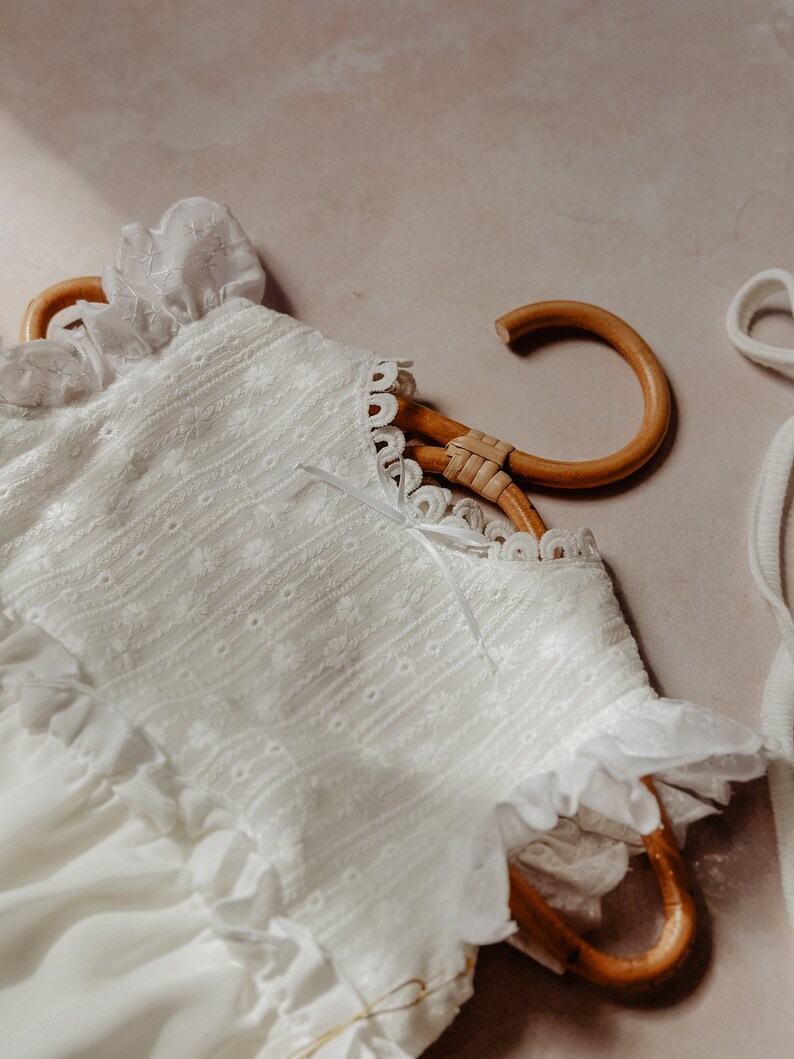 May include: A white baby dress with floral embroidery and ruffled trim hangs on a wooden hanger. The dress features a delicate lace collar and a small bow. The soft, light-coloured fabric suggests a vintage or heirloom style.