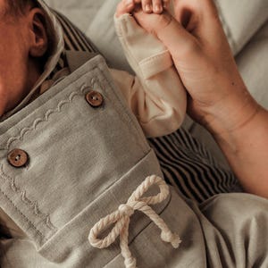 Op de afbeelding: Een lichtgrijze baby-outfit met een capuchon, houten knopen en een decoratieve touwstrik. De outfit is gemaakt van zachte stof en heeft een geschulpte rand. Een kleine hand wordt vastgehouden door een volwassen hand.