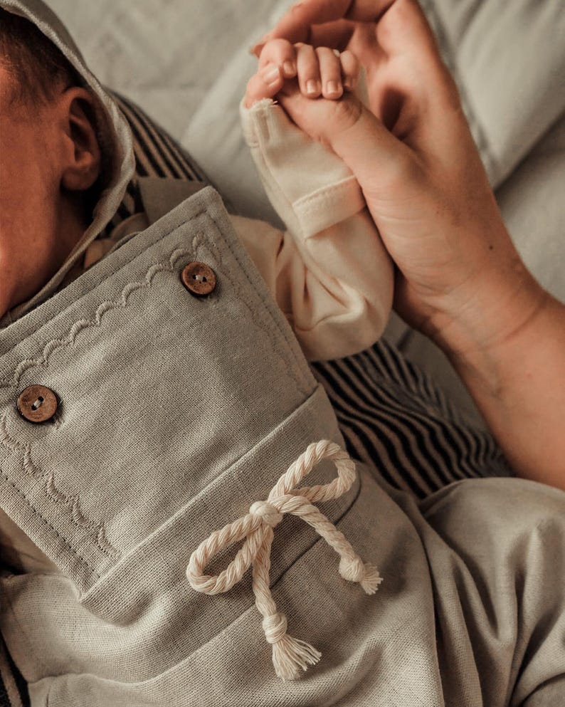 Op de afbeelding: Een lichtgrijze baby-outfit met een capuchon, houten knopen en een decoratieve touwstrik. De outfit is gemaakt van zachte stof en heeft een geschulpte rand. Een kleine hand wordt vastgehouden door een volwassen hand.