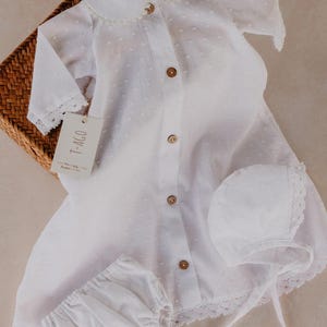 以下が含まれることがあります： 白のベビー服セット。ワンピース、ボンネット、ブルマが含まれています。ワンピースは襟、木製ボタン、レーストリムが特徴です。セットは籐かごの中にあります。「T-AGO」と書かれたタグが付いています。