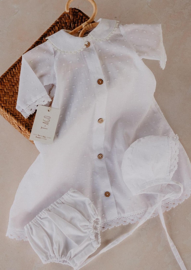 以下が含まれることがあります： 白のベビー服セット。ワンピース、ボンネット、ブルマが含まれています。ワンピースは襟、木製ボタン、レーストリムが特徴です。セットは籐かごの中にあります。「T-AGO」と書かれたタグが付いています。