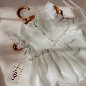 May include: A white baby romper with eyelet detailing and ruffle trim, displayed on a bamboo hanger. The romper features a button-up front and a flowing skirt. A tag with the brand name "tago" is attached.