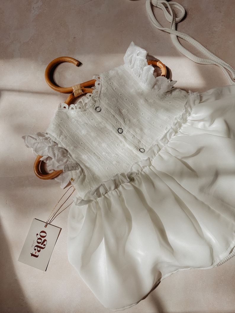 May include: A white baby romper with eyelet detailing and ruffle trim, displayed on a bamboo hanger. The romper features a button-up front and a flowing skirt. A tag with the brand name "tago" is attached.