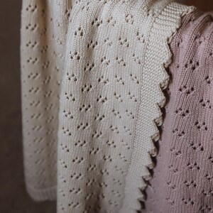 May include: Cream-colored knitted blanket with a delicate openwork pattern, draped over a chair. The blanket has a scalloped edge and is paired with a pink knitted garment, creating a soft, textured aesthetic.