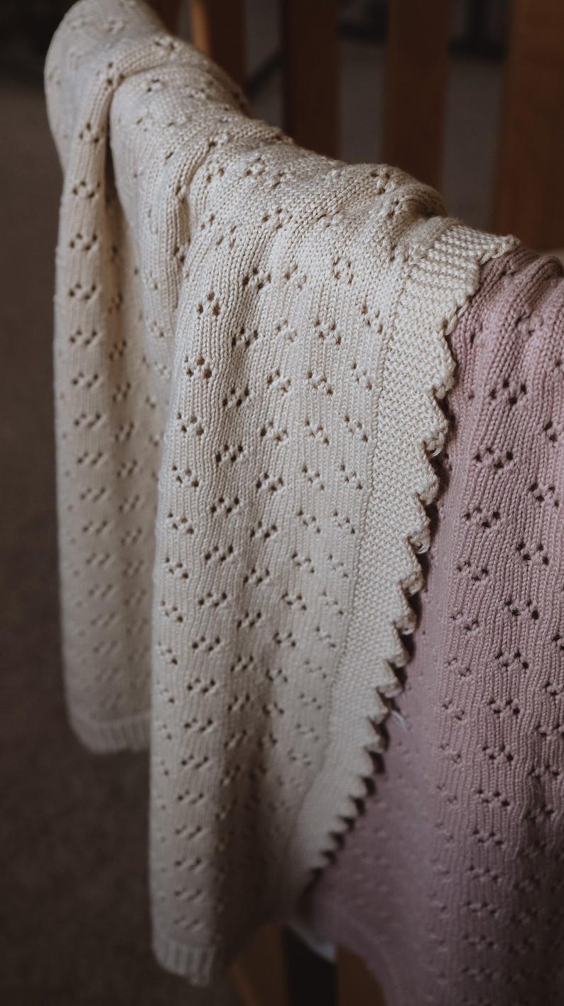 May include: Cream-colored knitted blanket with a delicate openwork pattern, draped over a chair. The blanket has a scalloped edge and is paired with a pink knitted garment, creating a soft, textured aesthetic.
