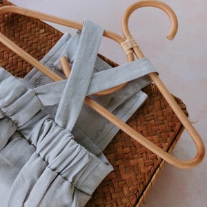 Op de afbeelding: Een lichtgrijze romper met een kruisrugontwerp, weergegeven op een houten kleerhanger. De romper is gemaakt van zachte stof en rust op een geweven mand, wat een minimalistische esthetiek cre&euml;ert. De elastische tailleband en bandjes van de romper zijn zichtbaar.