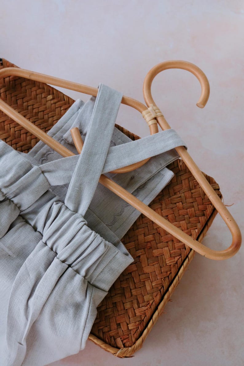 Op de afbeelding: Een lichtgrijze romper met een kruisrugontwerp, weergegeven op een houten kleerhanger. De romper is gemaakt van zachte stof en rust op een geweven mand, wat een minimalistische esthetiek cre&euml;ert. De elastische tailleband en bandjes van de romper zijn zichtbaar.