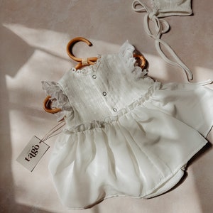 May include: A white baby dress and bonnet set. The dress has a ruffled bodice, eyelet detailing, and a flowing skirt. The bonnet features a lace trim and ties. The dress is on a wooden hanger, with a tag that reads "tago".
