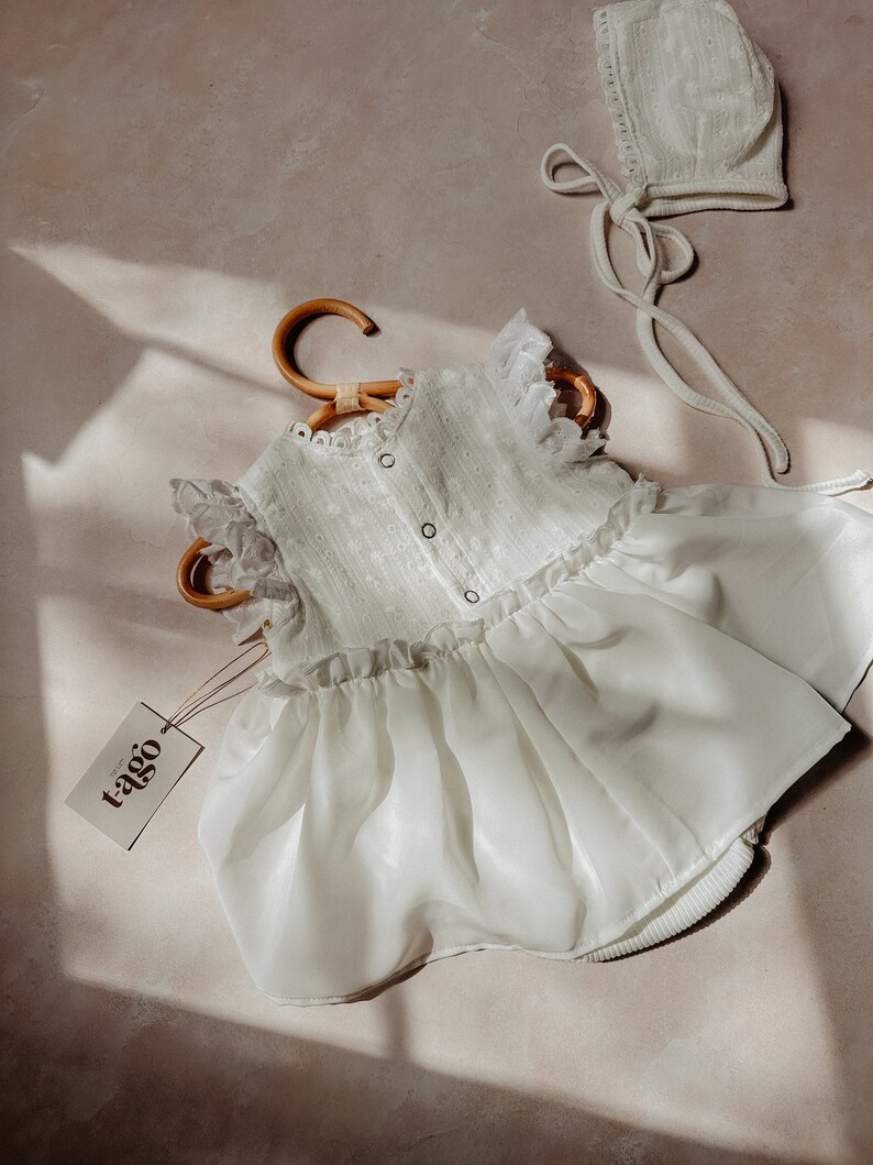 May include: A white baby dress and bonnet set. The dress has a ruffled bodice, eyelet detailing, and a flowing skirt. The bonnet features a lace trim and ties. The dress is on a wooden hanger, with a tag that reads "tago".