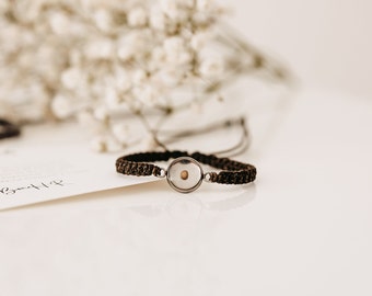 May include: A brown braided bracelet with a silver circular pendant containing a small seed. The bracelet is lying on a white surface.