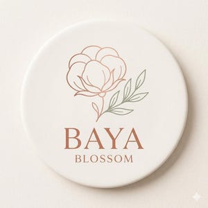 Op de afbeelding: Een ronde, crèmekleurige onderzetter met een bloemmotief en de tekst "BAYA BLOSSOM". Het ontwerp toont een roségouden omtrek van een katoenbloem en groene bladeren. De tekst is in een warme bruine kleur.