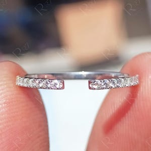 Może przedstawiać: Srebrny pierścionek o rozdzielonej konstrukcji, z rzędem małych, przezroczystych kamieni szlachetnych osadzonych wzdłuż górnej połowy obrączki. Pierścionek trzymany jest między dwoma palcami, prezentując jego delikatne detale i błyszczący wygląd.