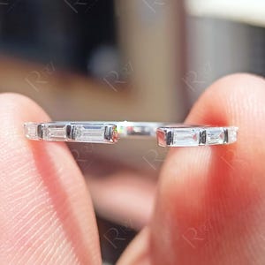 Puede incluir: Un anillo abierto de tono plateado que presenta una serie de piedras transparentes de corte baguette rectangular engastadas a lo largo de la banda. El anillo tiene una abertura en la parte superior, mostrando un acabado metálico y piedras alineadas.