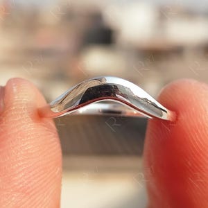 May include: A close-up of a silver ring with a curved design. The ring is held between two fingers, showcasing its polished, reflective surface. The ring's shape is a smooth, gentle curve, reflecting the light.