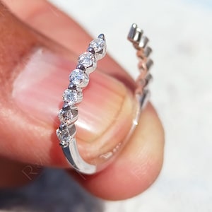 May include: A silver ring featuring a line of small, clear gemstones. The ring is held in a hand, highlighting the intricate design and sparkle of the stones. The open design of the ring gives it a contemporary feel.