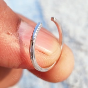 May include: A silver ring with an open design. The ring is held between two fingers and has a smooth, curved shape. The ends of the ring are slightly apart. The ring appears to be made of a shiny metal.
