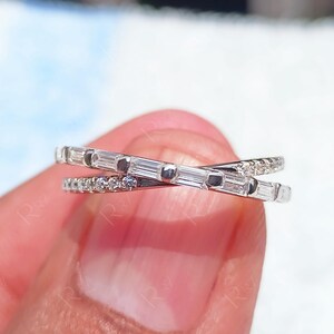 Op de afbeelding: Een zilverkleurige ring met een kruisontwerp. Eén band is bezet met kleine, ronde edelstenen, de andere met rechthoekige edelstenen. De ring wordt tussen vingers gehouden, wat de ingewikkelde details en elegante stijl benadrukt.