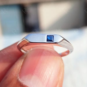 May include: A polished silver-toned metal ring featuring a flat, rectangular top surface. A single, small, square-cut blue gemstone is set flush into the center of the flat metal bar, creating a clean and simple design for this finger accessory.