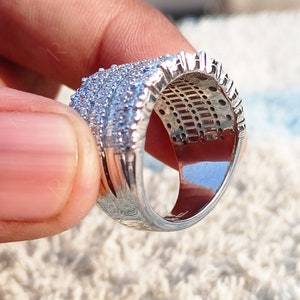 May include: A silver ring with a wide band and a row of small, clear stones. The ring is set on a white, fluffy surface.