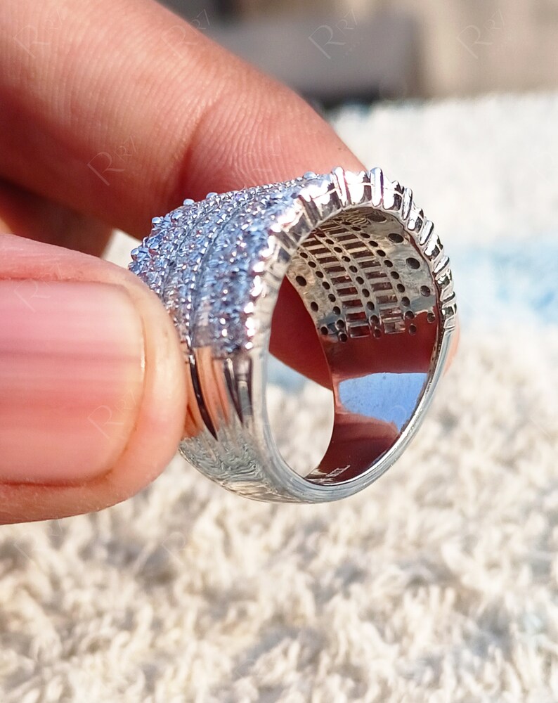 May include: A silver ring with a wide band and a row of small, clear stones. The ring is set on a white, fluffy surface.