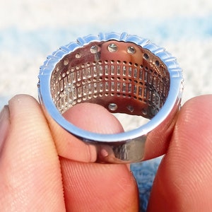 May include: A silver ring with a row of small, clear stones set in a band. The ring is held between two fingers.