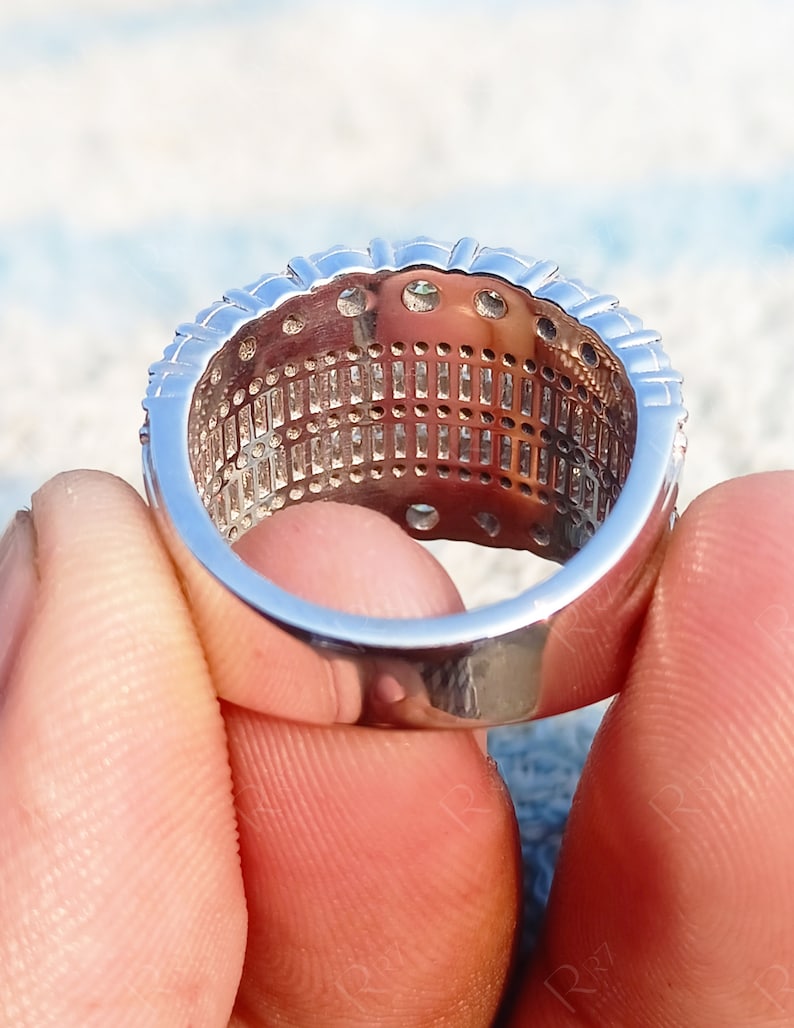 May include: A silver ring with a row of small, clear stones set in a band. The ring is held between two fingers.