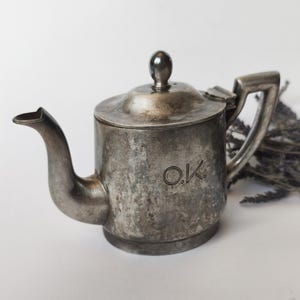 Op de afbeelding: Een theepot van aangeslagen zilverkleurig metaal met een gebogen tuit, een scharnierend deksel met een kleine bolvormige knop en een hoekig handvat. Op de zijkant van de theepot staan de letters O.K. gegraveerd in een eenvoudig, duidelijk lettertype.