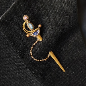 Könnte beinhalten: Goldfarbene, schwertförmige Brosche mit aufwendigen Mosaikdetails in Blau, Weiß und Rot. Die Brosche hat eine kleine Kette und eine Nadel. Der Hintergrund ist ein dunkler, strukturierter Stoff.