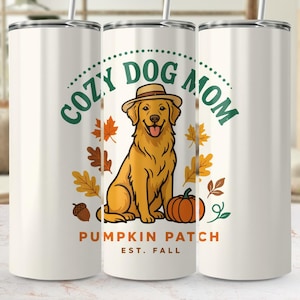 Peut inclure: Un gobelet cylindrique blanc avec une paille en métal illustré d'un golden retriever portant un chapeau, entouré de feuilles d'automne, d'un gland et d'une citrouille. Le texte indique COZY DOG MOM, PUMPKIN PATCH et EST. FALL en vert et orange.
