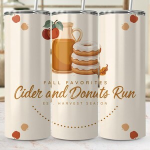 Könnte beinhalten: Ein zylindrischer Becher mit cremefarbenem Hintergrund, verziert mit einer Grafik aus einem Glaskrug mit Apfelwein, einem Stapel aus drei glasierten Donuts und roten Äpfeln. Der Text lautet FALL FAVORITES Cider and Donuts Run EST. HARVEST SEASON.