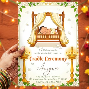 May include: An invitation card for a Cradle Ceremony, with an illustration of a baby in a cradle. The card includes the text "Cradle Ceremony of Aaryan" and event details. The card is held by a person with henna tattoos and colourful bangles.