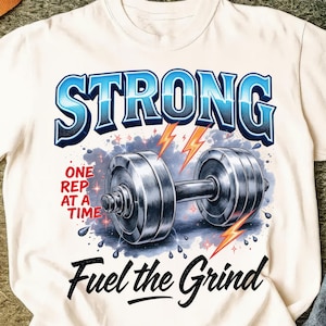 Può includere: T-shirt color crema con un design grafico con la scritta "STRONG" in blu, un manubrio, fulmini e le frasi "ONE REP AT A TIME" e "Fuel the Grind".