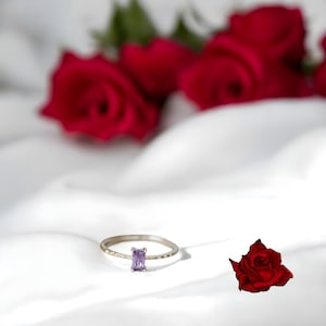 Peut inclure: Une délicate bague en argent avec une pierre précieuse violette sur un fond blanc avec des roses rouges.