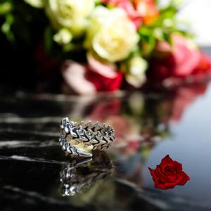 Peut inclure: Une bague en argent avec un design texturé unique ressemblant à l'épine dorsale d'un dragon. La bague est posée sur une surface noire avec un arrière-plan flou de roses rouges et blanches.