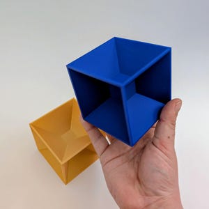 May include: A hand holds a bright blue, 3D-printed cube with an open top and a square base. A yellow cube is next to it. The cubes have a geometric design and are likely used for display or storage.