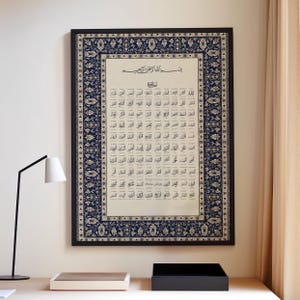Op de afbeelding: Ingelijst kunstwerk met Arabische kalligrafie op een crèmekleurige achtergrond, omgeven door een marineblauwe en crèmekleurige bloemenrand. De lijst is zwart. Het kunstwerk wordt tentoongesteld op een lichtgekleurde muur.