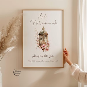 May include: A framed print with the words "Eid Mubarak" in elegant script. The artwork features a detailed illustration of a lantern surrounded by pink flowers. The bottom of the print includes Arabic text and the English translation, "May Allah accept it from us and from you."