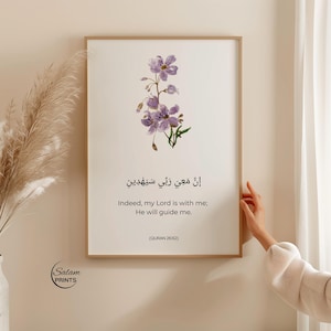 Puede incluir: Un cuadro enmarcado con una ilustración de flores moradas. El cuadro incluye texto en árabe y en inglés: «En verdad, mi Señor está conmigo; Él me guiará». El cuadro es de Salam Prints.