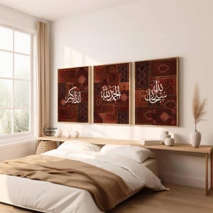 Op de afbeelding: Drie ingelijste wandkunstwerken met Arabische kalligrafie op een patroon, roodbruine achtergrond. De frames zijn licht van kleur. Het kunstwerk wordt tentoongesteld boven een bed met witte lakens en een bruine deken.
