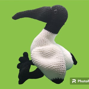 May include: A crocheted stuffed bird with a white body and black head, beak, and legs. The bird is sitting on a green background.