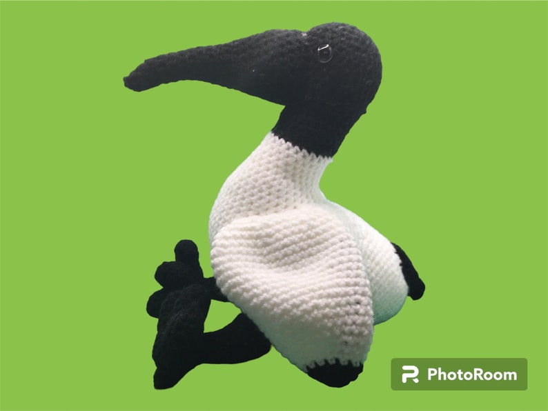 May include: A crocheted stuffed bird with a white body and black head, beak, and legs. The bird is sitting on a green background.