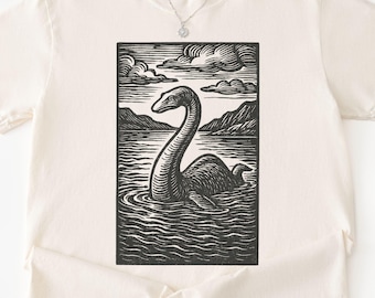 May include: Cream-colored t-shirt featuring a black and white illustration of the Loch Ness Monster in a lake scene. The design includes mountains, clouds, and water ripples. The shirt has short sleeves and a crew neck.