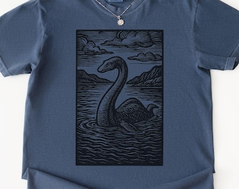 May include: A navy blue t-shirt featuring a black and white illustration of a Loch Ness Monster in a lake with mountains and clouds. The shirt has short sleeves and a crew neck. A silver necklace with a flower pendant is visible.