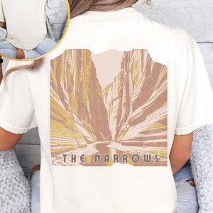 May include: A white t-shirt with a graphic of a canyon scene in shades of brown and yellow. The text "THE NARROWS" is printed below the image.